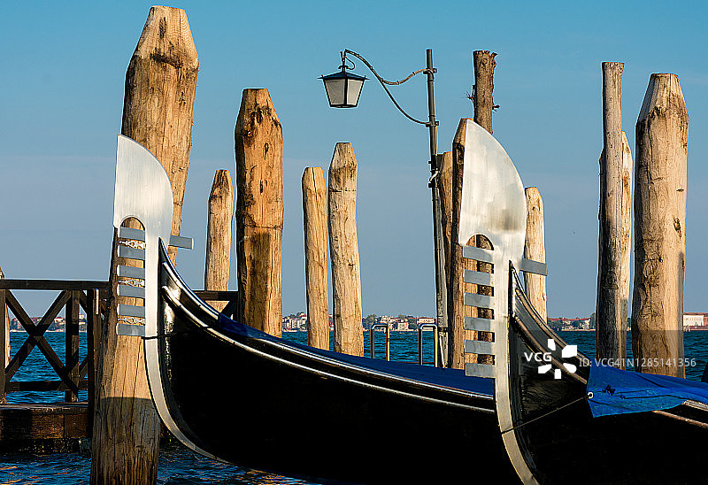 日落时分，两艘意大利贡多拉系在码头的柱子上，漂浮在蓝色湖面上。照片仅显示贡多拉的船头图片素材