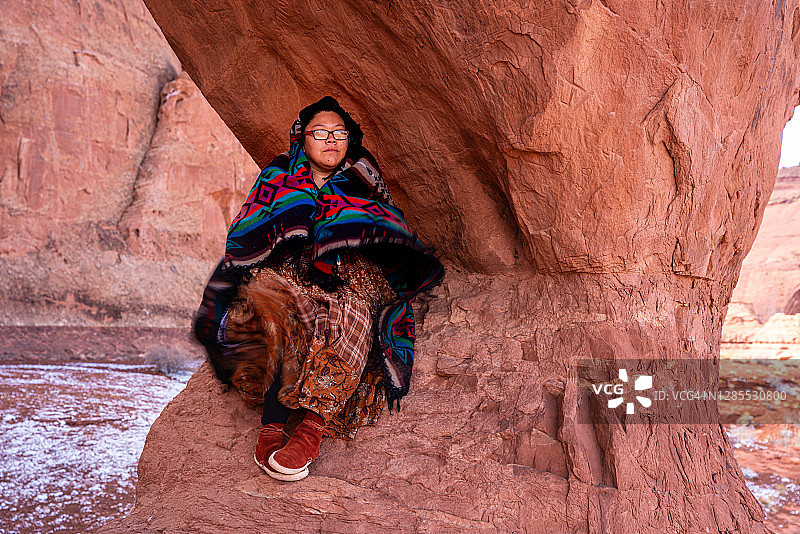 一位中国台湾纳瓦霍族年轻女子裹着传统的纳瓦霍羊毛毯，坐在犹他州纪念碑谷的标志性泪滴拱门中图片素材