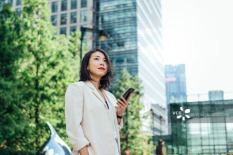 自信的年轻女商人站在金融区的商业建筑前使用智能手机图片素材