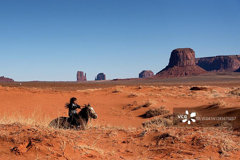 在驼峰山附近的沙漠中骑马的年轻纳瓦霍男孩，背景是纪念碑谷图片素材