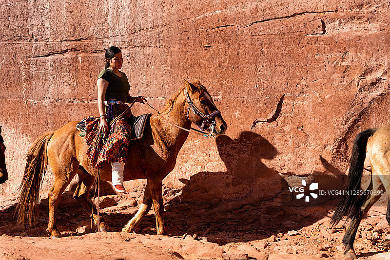 一位年轻的纳瓦霍族妇女在刻满岩画的巨大石墙附近骑马图片素材