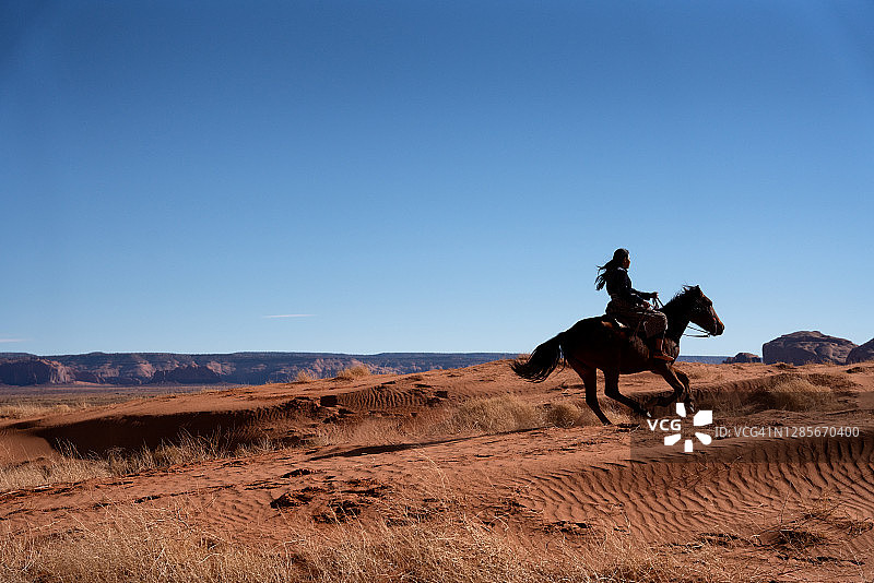 一位穿着传统服装的年轻纳瓦霍族妇女骑着马穿过纪念碑谷骆驼峰附近的沙漠，亚利桑那州图片素材