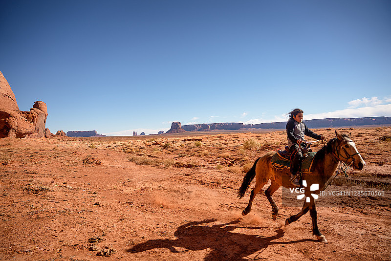 在驼峰山附近的沙漠中骑马的年轻纳瓦霍男孩，背景是纪念碑谷图片素材