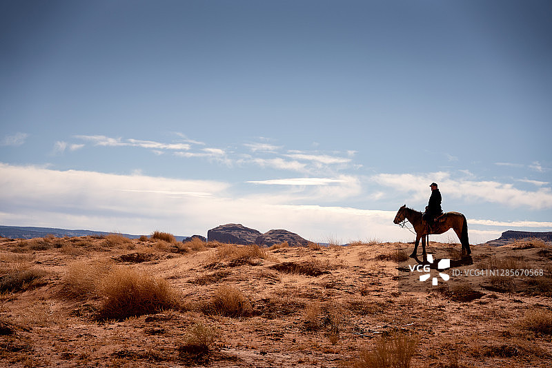 年长的西班牙裔男人在亚利桑那州北部纪念碑谷部落公园附近的崎岖沙漠中骑马图片素材