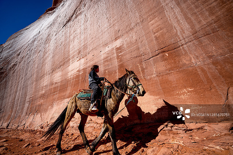 一位年轻的纳瓦霍族男孩在刻满岩画的巨石墙附近骑马图片素材