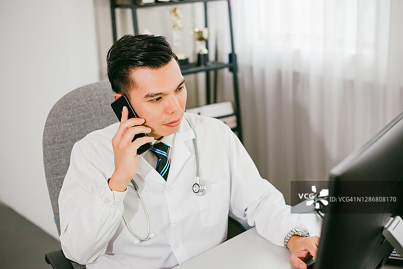 亚洲外科医生在办公室阅读电脑上的医疗报告并使用智能手机图片素材