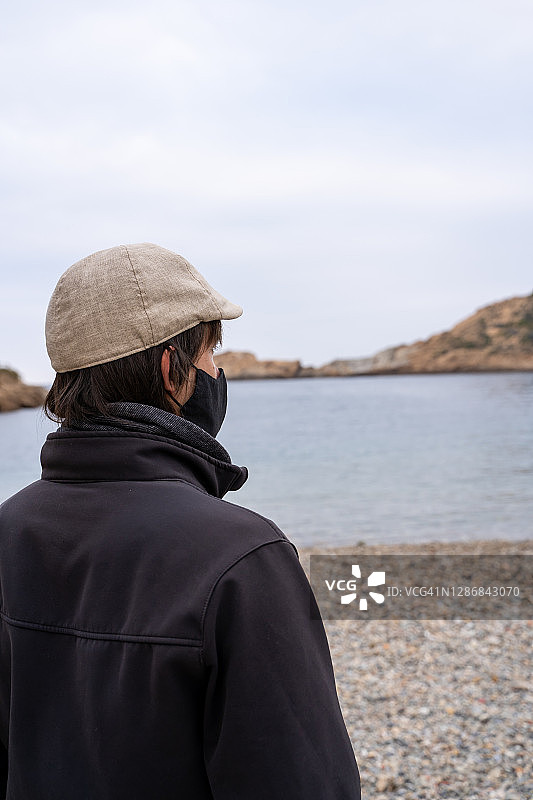 戴贝雷帽和口罩的男人面朝大海站立的竖版照片，西班牙加泰罗尼亚Sa Tuna海滩图片素材