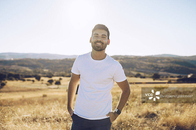 晴天里，微笑的年轻男人站在田野上，双手插在口袋里图片素材