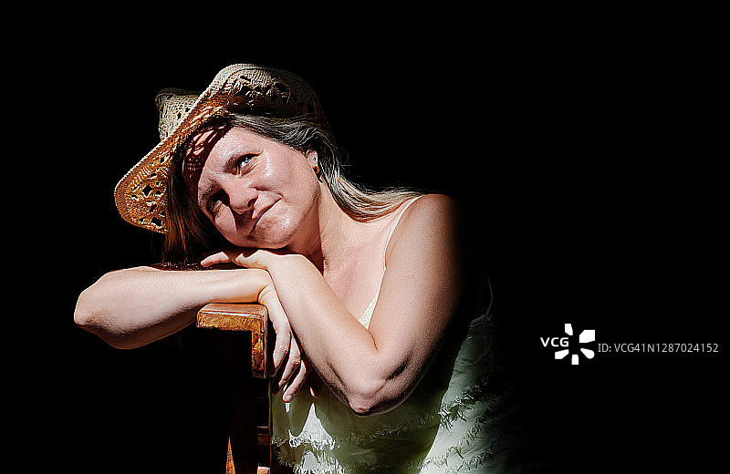 一位戴着草帽，面带微笑，将头靠在木桌上的40-50岁金发女人的肖像正面图。该女子在黑色背景下看着镜头。水平构图图片素材