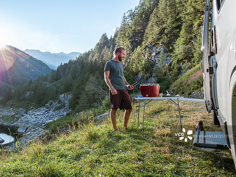 在面包车外用便携式烧烤炉做饭的男人，山景图片素材