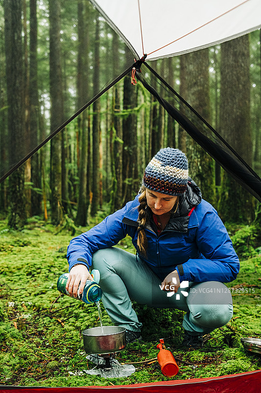 成年女人在茂密的热带雨林中，将瓶子里的水倒进野营炉上的锅里做饭图片素材