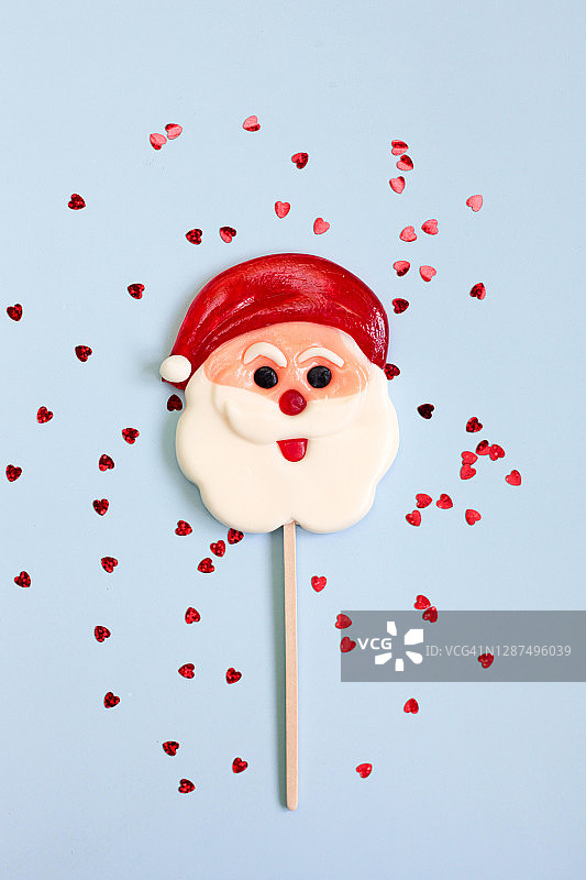 圣诞节贺卡：圣诞老人造型棒棒糖和红色发光纸屑，蓝色粉彩背景，竖版图片素材