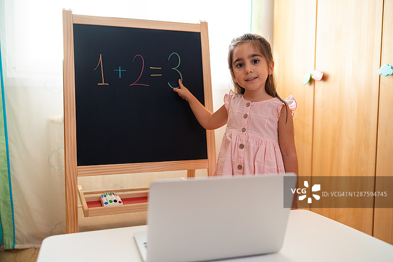 在家用笔记本电脑上网课的小女学生，展示她在黑板上写的加法算式图片素材