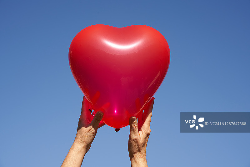 两只手拿着红色心形气球，象征着健康的心脏、心脏移植或情人节，蓝色天空背景。图片素材