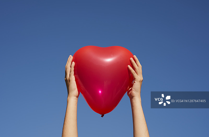 两只手拿着红色心形气球，象征着健康的心脏、心脏移植或情人节，蓝色天空背景。图片素材