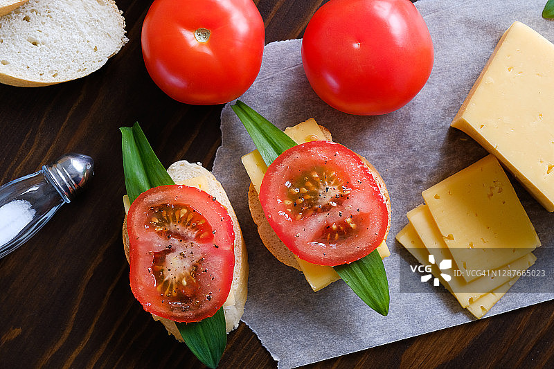 木桌上的面包片、三明治、刀、砧板、红番茄、盐瓶、奶酪和蔬菜特写。烹饪早餐或零食，有机农场产品，居家生活图片素材