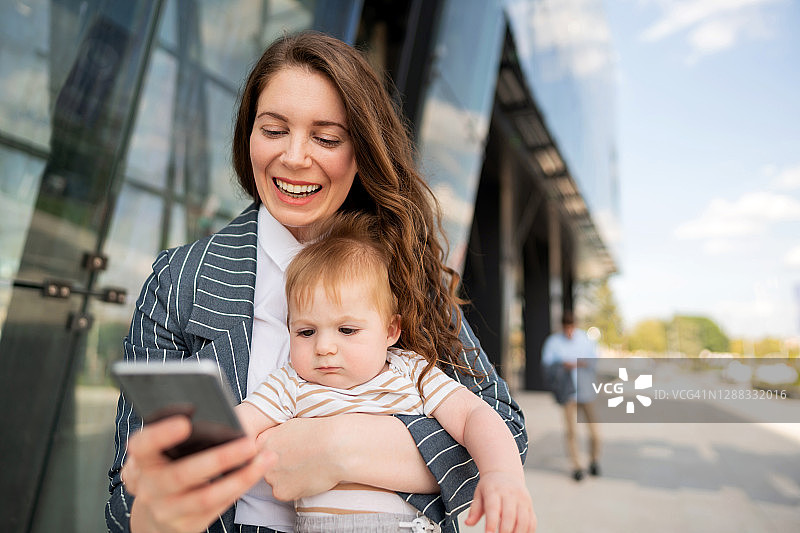 微笑的母亲抱着她可爱的男婴，同时拿着智能手机发短信图片素材