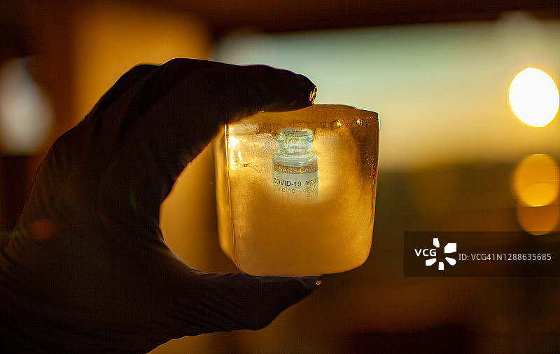 戴着外科手套的手拿着冰块，里面装着低温储存的COVID-19疫苗瓶，标签为SARS-CoV-2对抗冠状病毒图片素材