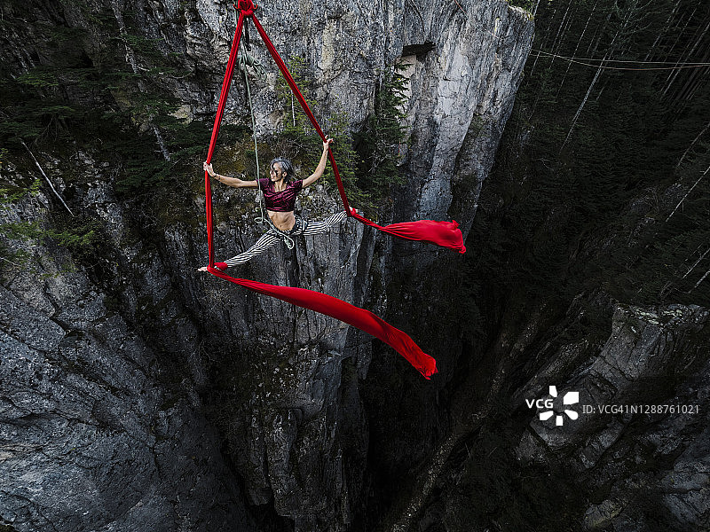 一位女性杂技演员在红色空中绸缎上表演劈叉，绸缎悬挂在高高的岩石峡谷面上图片素材