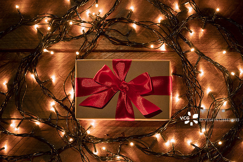 牛皮纸包装的惊喜礼物，用鲜红的丝带系着，在木桌上，背景是金色闪烁的童话灯串。在节日的前夜给予和赠送礼物。快乐圣诞节的概念图片素材