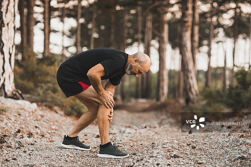 在森林户外跑步时膝盖受伤：一位老人用手扶着膝盖，感到疼痛。韧带扭伤或半月板问题。图片素材