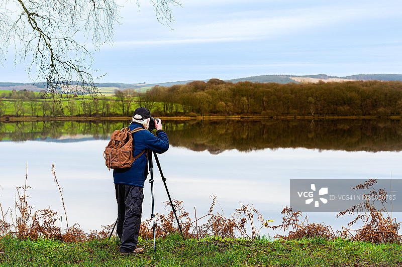 秋日清晨，一位年长的男人在拍摄平静的苏格兰湖泊图片素材