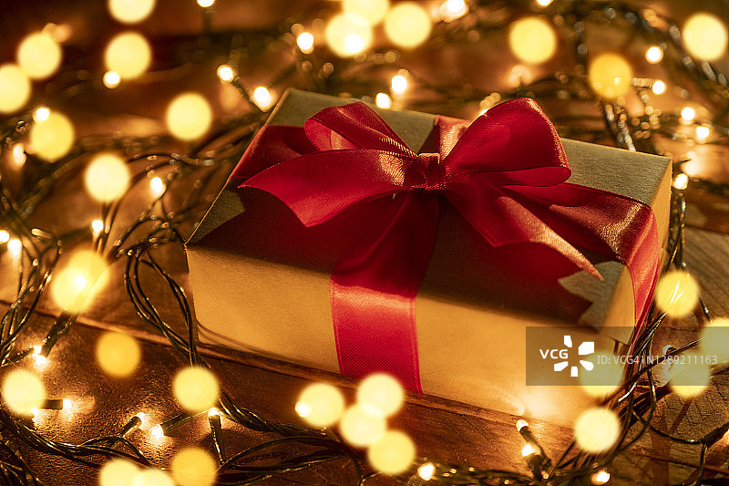 牛皮纸包装的惊喜礼物，用鲜红的丝带系着，在木桌上，背景是金色闪烁的童话灯串。在节日的前夜给予和赠送礼物。快乐圣诞节的概念图片素材