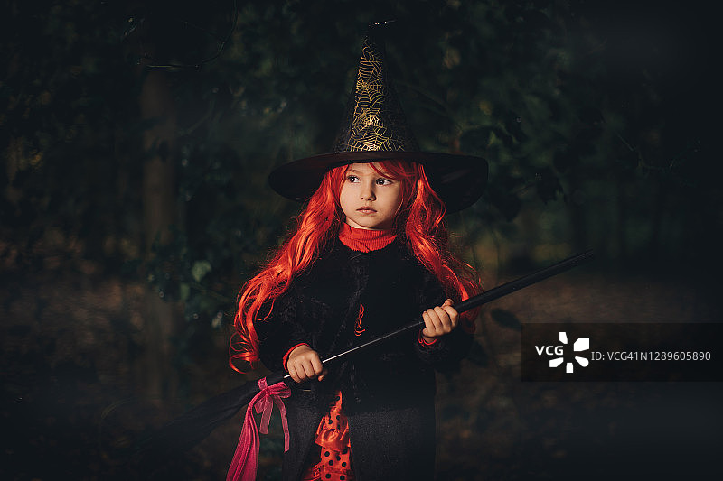 红发万圣节女巫装扮的小女孩在黑暗的森林里拿着扫帚图片素材