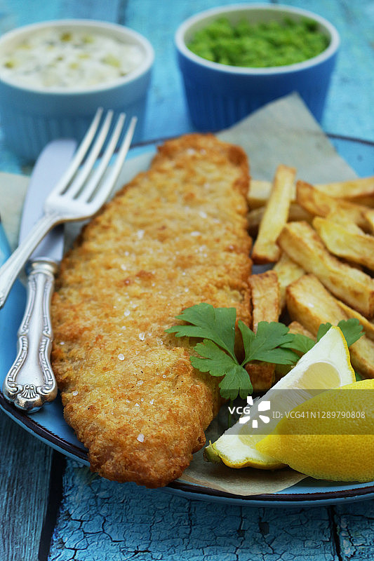 炸鱼薯条（鳕鱼）特写，用欧芹装饰，放在绿松石盘子上，配有防油纸、刀叉、柠檬片、鞑靼酱和豌豆泥，绿松石木纹背景，焦点在前图片素材
