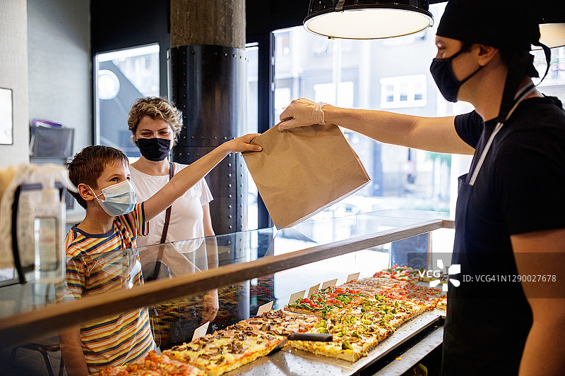 戴防护口罩的男孩在披萨店员工处隔着玻璃柜台取餐图片素材