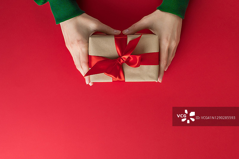 身穿绿色毛衣的女孩拿着一个漂亮的盒子，盒子上系着一条鲜艳的丝带，背景是红色。女人在节日里打开礼物。快乐圣诞节的概念图片素材
