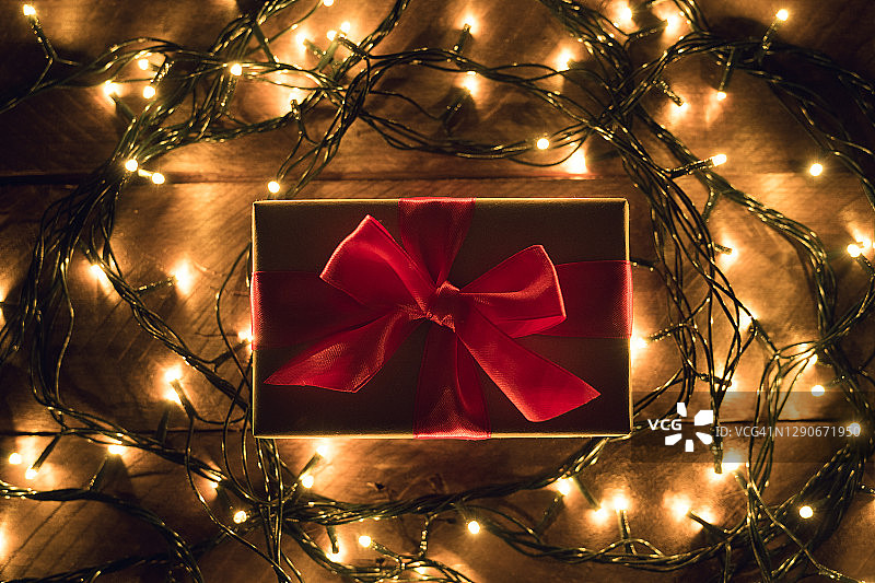 牛皮纸包装的惊喜礼物，用鲜红的丝带系着，在木桌上，背景是金色闪烁的童话灯串。在节日的前夜给予和赠送礼物。快乐圣诞节的概念图片素材