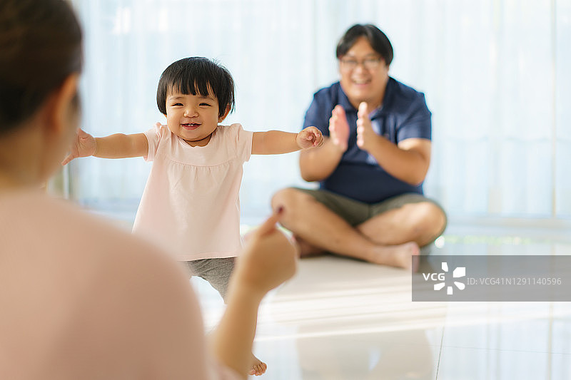 亚洲女婴迈出人生的第一步，走向她的母亲。快乐的小宝宝在父亲的帮助和教导下，学会在家里轻轻地走路图片素材