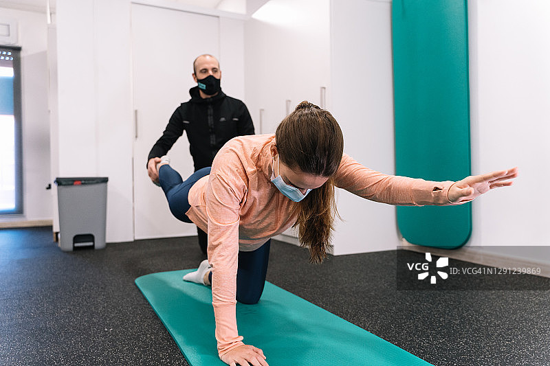 在健身房里，教练帮助一位戴着面罩在垫子上做伸展运动的年轻女子图片素材