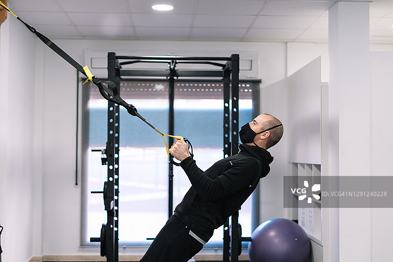 在健身房用TRX悬挂带做悬吊运动的戴口罩男人图片素材