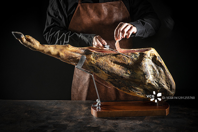 在阴暗背景下手持刀切割伊比利亚火腿的男人图片素材