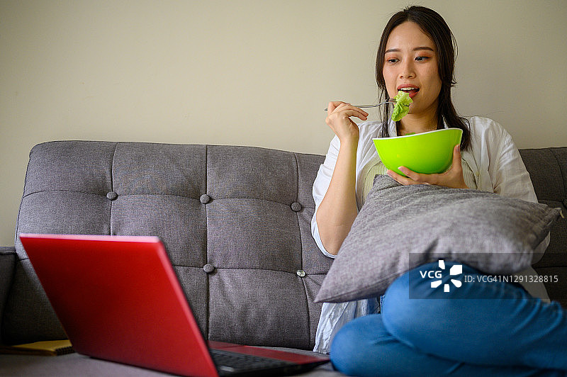 年轻漂亮的亚洲女子在客厅里吃健康食品并使用笔记本电脑图片素材