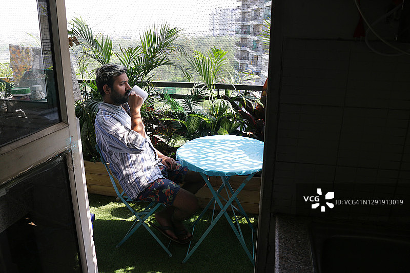 男子坐在阳台蓝色圆桌旁，沐浴阳光，手捧热咖啡，背景是热带丛林花园图片素材
