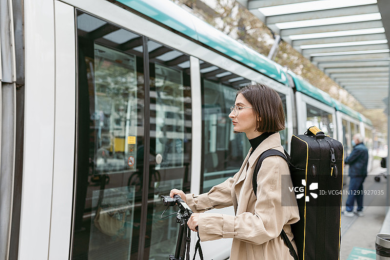 手提乐器箱、骑电动滑板车的女人在城市里等有轨电车图片素材