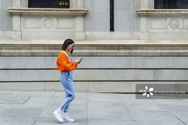 年轻女人在人行道上靠墙行走时使用手机图片素材