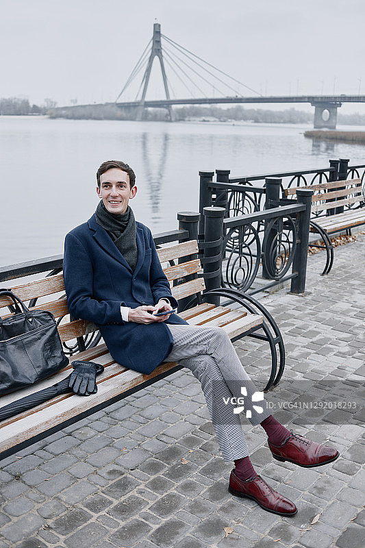 身穿经典外套坐在长椅上，手拿智能手机的男人图片素材