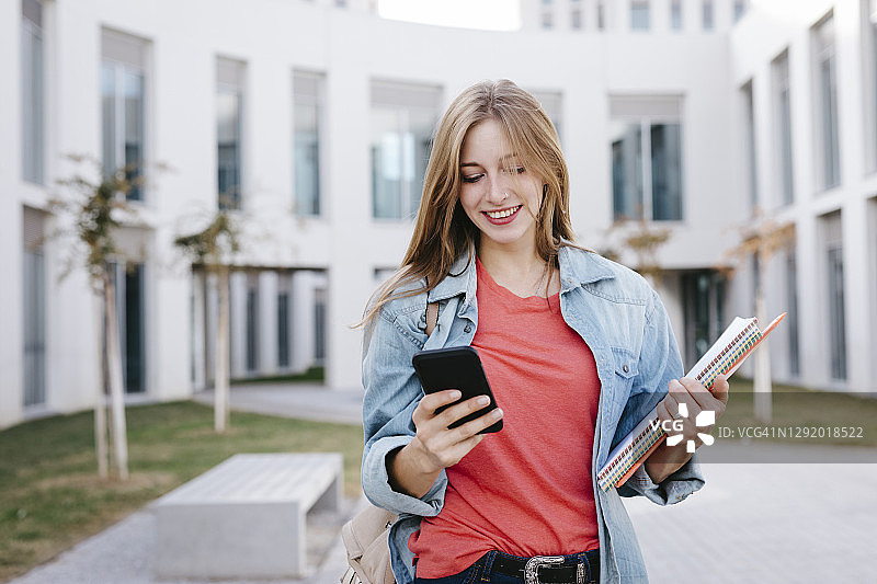 笑容灿烂的年轻金发女学生在大学使用智能手机图片素材