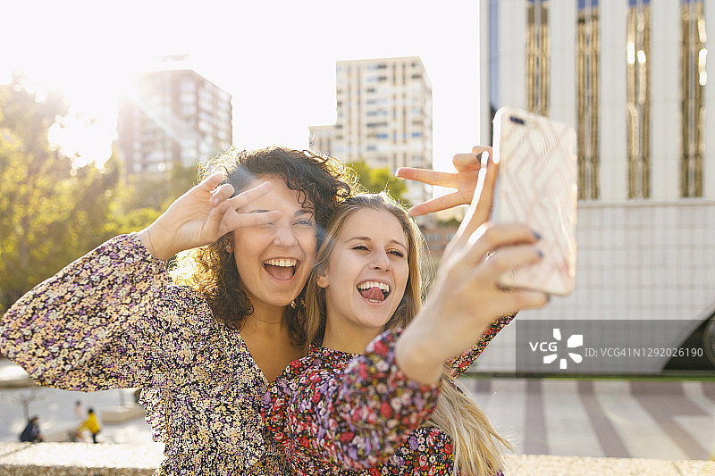 快乐的女性朋友在城市里用智能手机自拍时做出和平手势图片素材