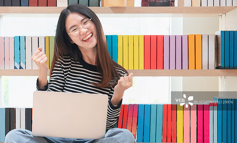 用笔记本电脑庆祝的亚洲女孩，成功的快乐姿势。学习和教育理念。女大学生在图书馆里拿着笔记本电脑高兴地笑着图片素材