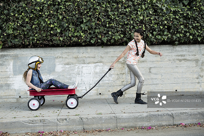 在人行道上拉着坐在四轮车里的朋友的快乐女孩图片素材