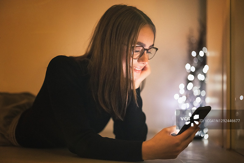 微笑的女人晚上在客厅地板上放松，查看她的手机图片素材