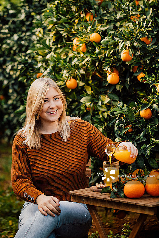 年轻女子将自家收获的鲜榨橙汁倒入玻璃杯中，对着镜头可爱地笑着图片素材