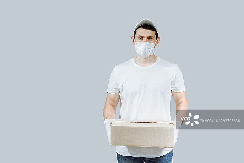 戴白色帽子、穿白色T恤制服、戴口罩手套的送货员拿着空纸板箱，灰色背景演播室服务、隔离、疫情、冠状病毒概念图片素材