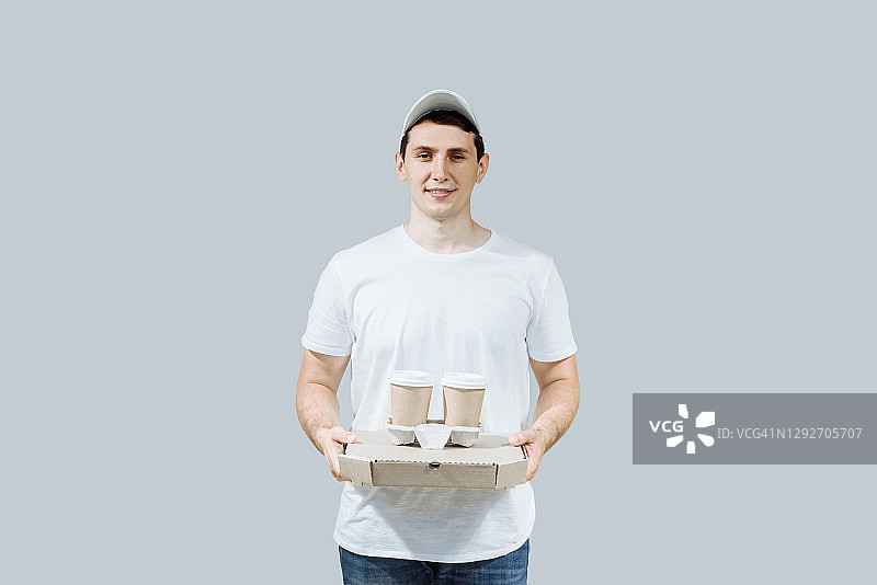这是您的披萨！快乐的年轻送货员拿着披萨盒和咖啡，在灰色背景下隔离。快递员有复制空间图片素材