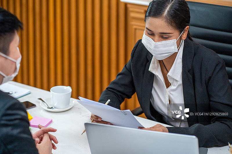 亚洲女工和男商务工人在联合办公空间一起使用笔记本电脑交谈，戴着防护口罩，防止PM2.5和新冠病毒。健康和团队合作理念图片素材
